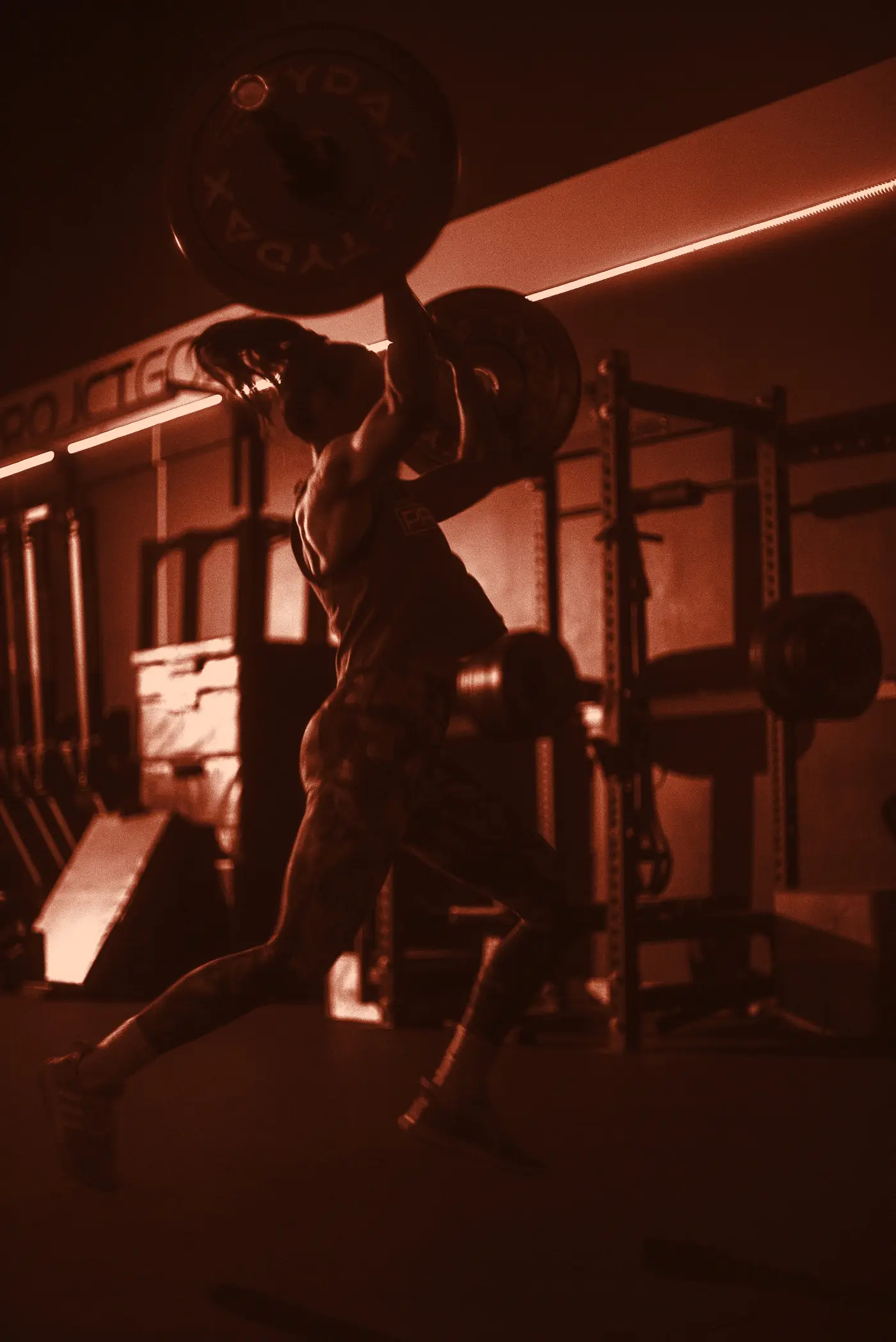 A lifter driving a loaded barbell overhead in a dark gym, lit orange